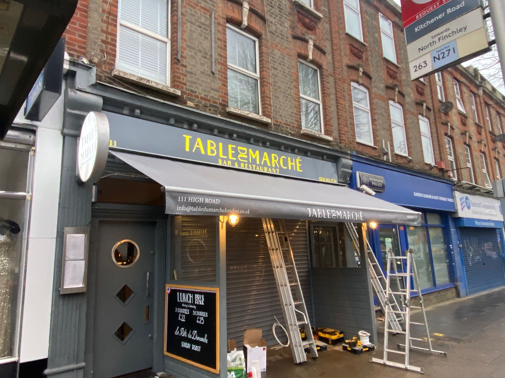 Angled view of Table du Marché showing the fitted grey sign tray, gold lettering and awning during installation.